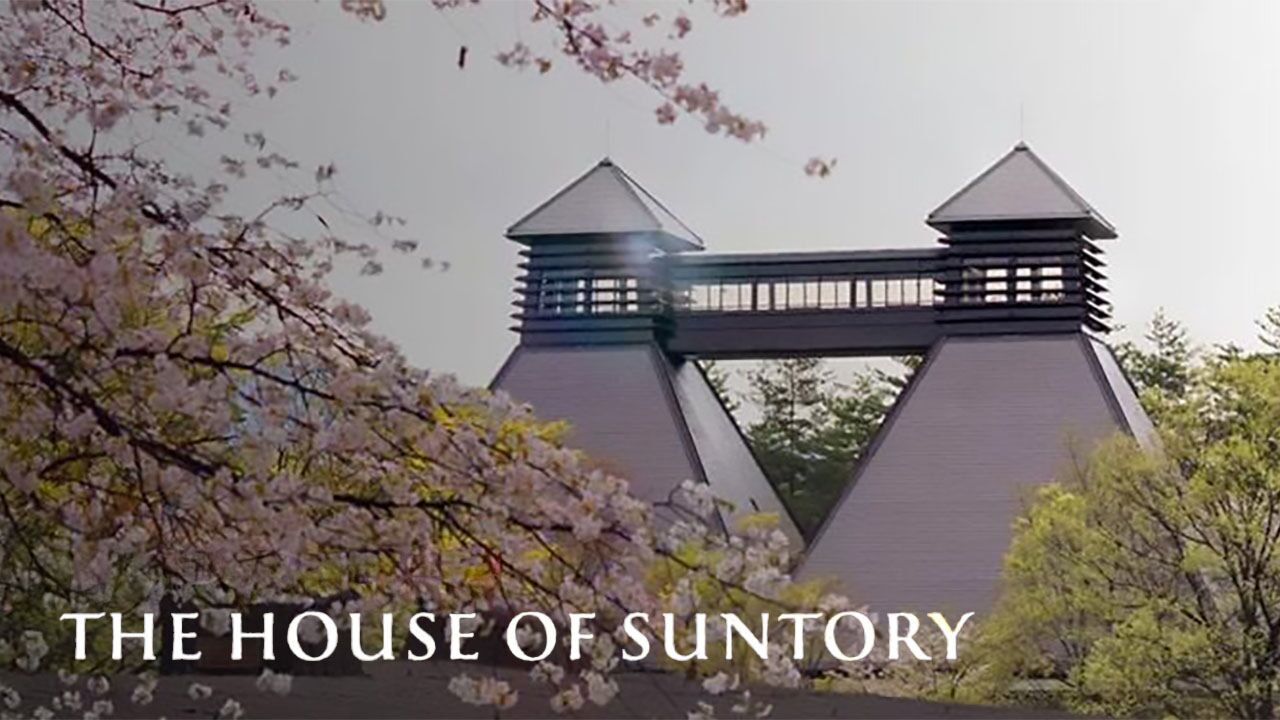 A photograph of the Suntory Japanese Whisky Distillery surrounded by Cherry Blossom and other trees. Superimposed in the bottom left corner there's a line of text reading: The House of Suntory