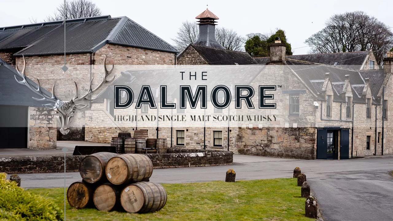 A photograph of The Dalmore distillery exterior; stone farmhouse-style building and casks on a grass patch. Superimposed text reads: The Dalmore Highland Single Malt Scotch Whisky.