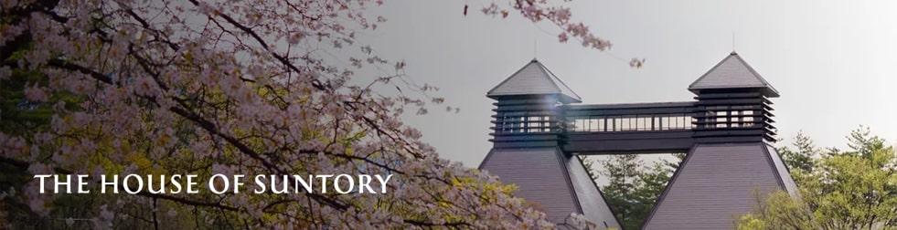 A photograph of the Suntory Japanese Whisky Distillery surrounded by Cherry Blossom and other trees. Superimposed in the bottom left corner there's a line of text reading: The House of Suntory